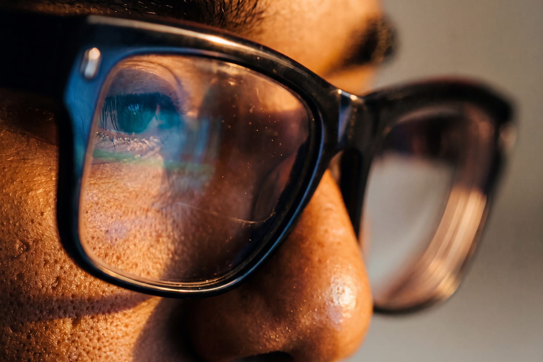 Extreme close-up of a person's eye behind thick black-framed glasses with screen light reflected in the lens.
