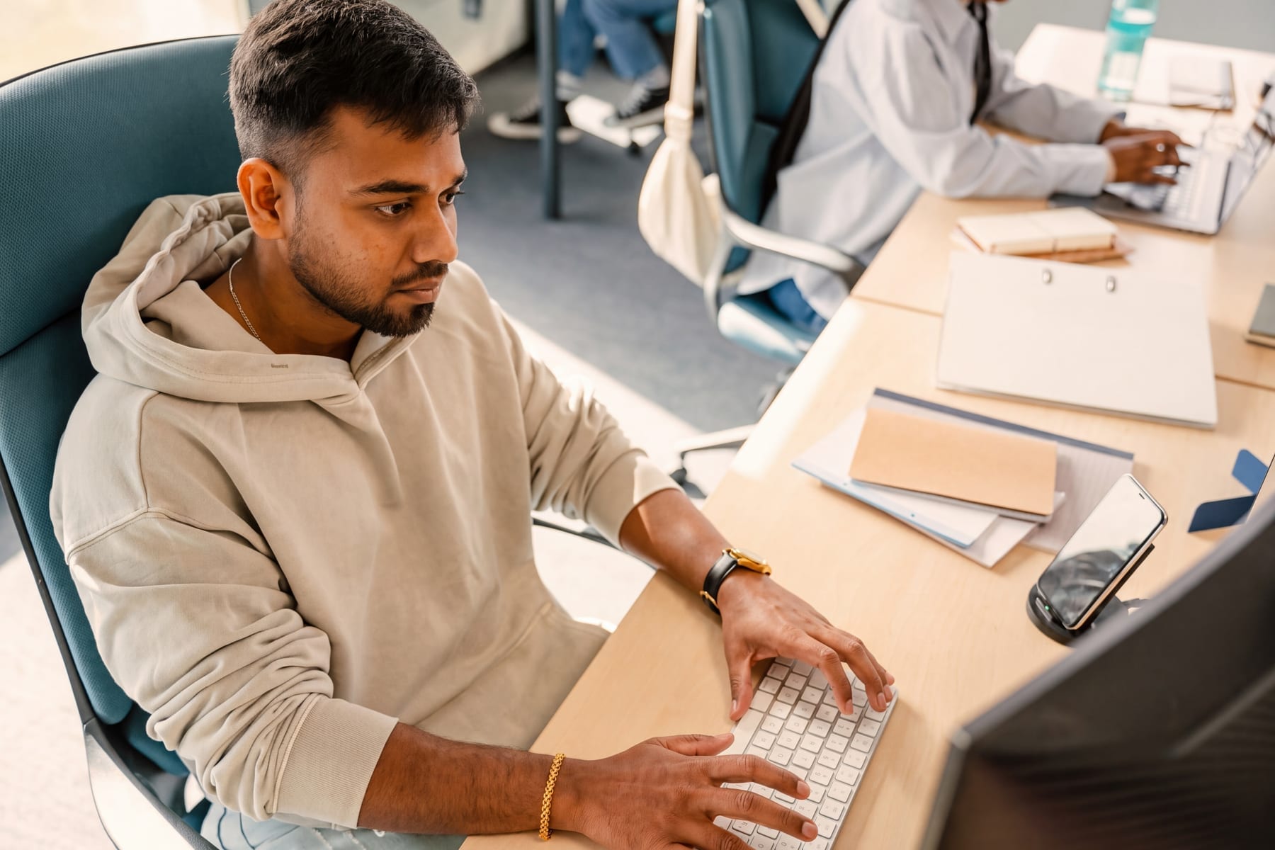 A young man in a beige hoodie types at a keyboard in an open-plan office while a coworker works at a shared desk behind him.