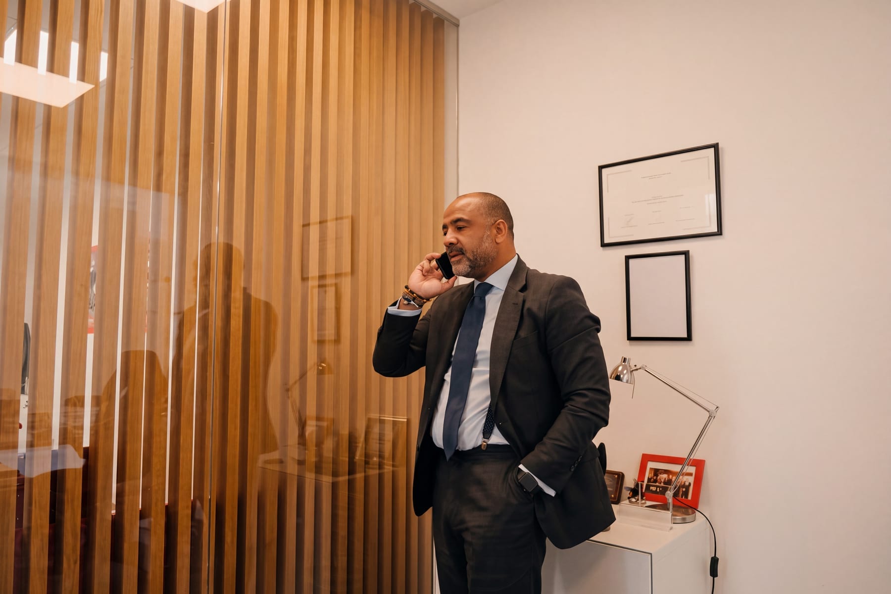 A professional man in a dark suit holds a phone to his ear in an office setting beside framed certificates.