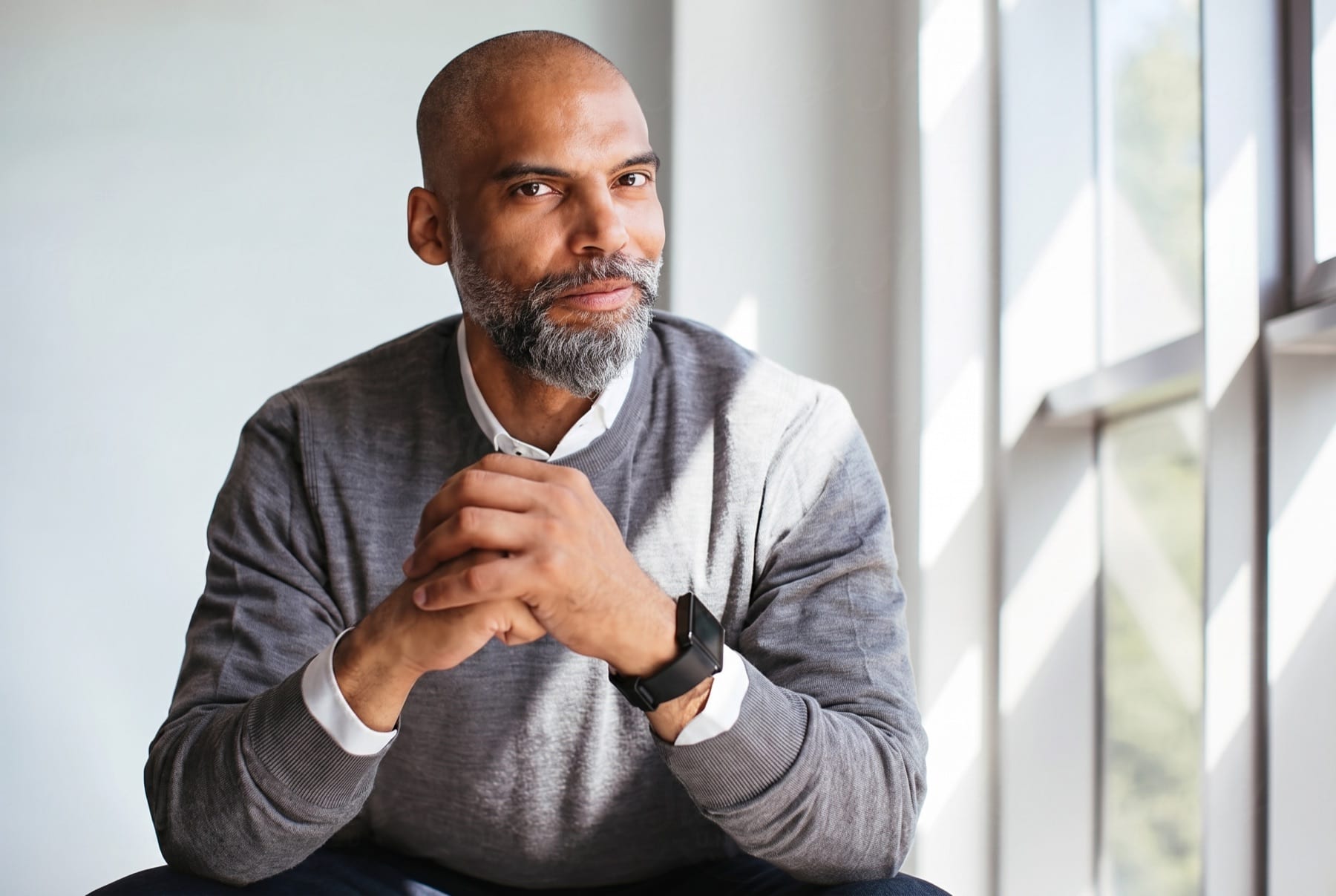 A man sits by a window with his hands clasped, gazing outward in contemplation.