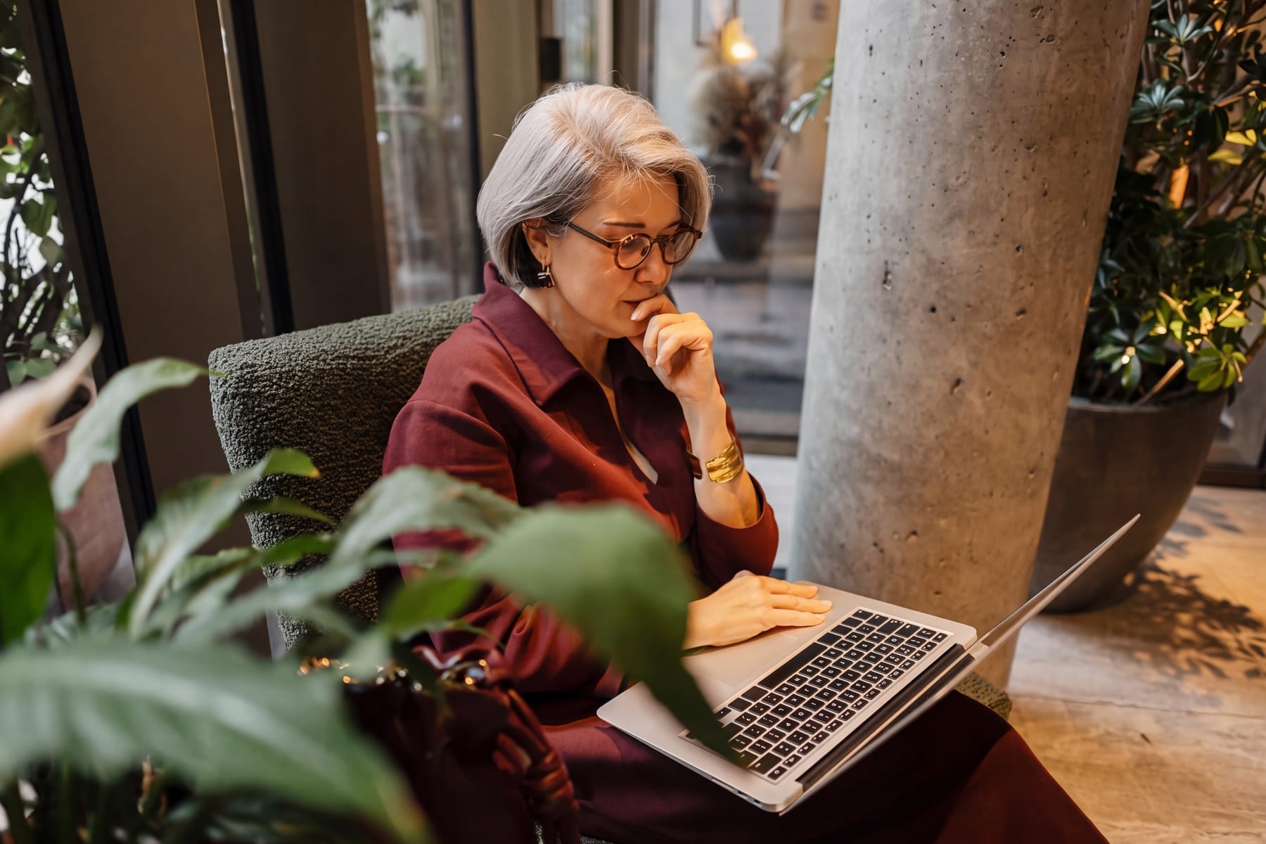 Woman in burgundy suit and glasses studies a laptop screen intently from an armchair, hand raised thoughtfully to her chin.